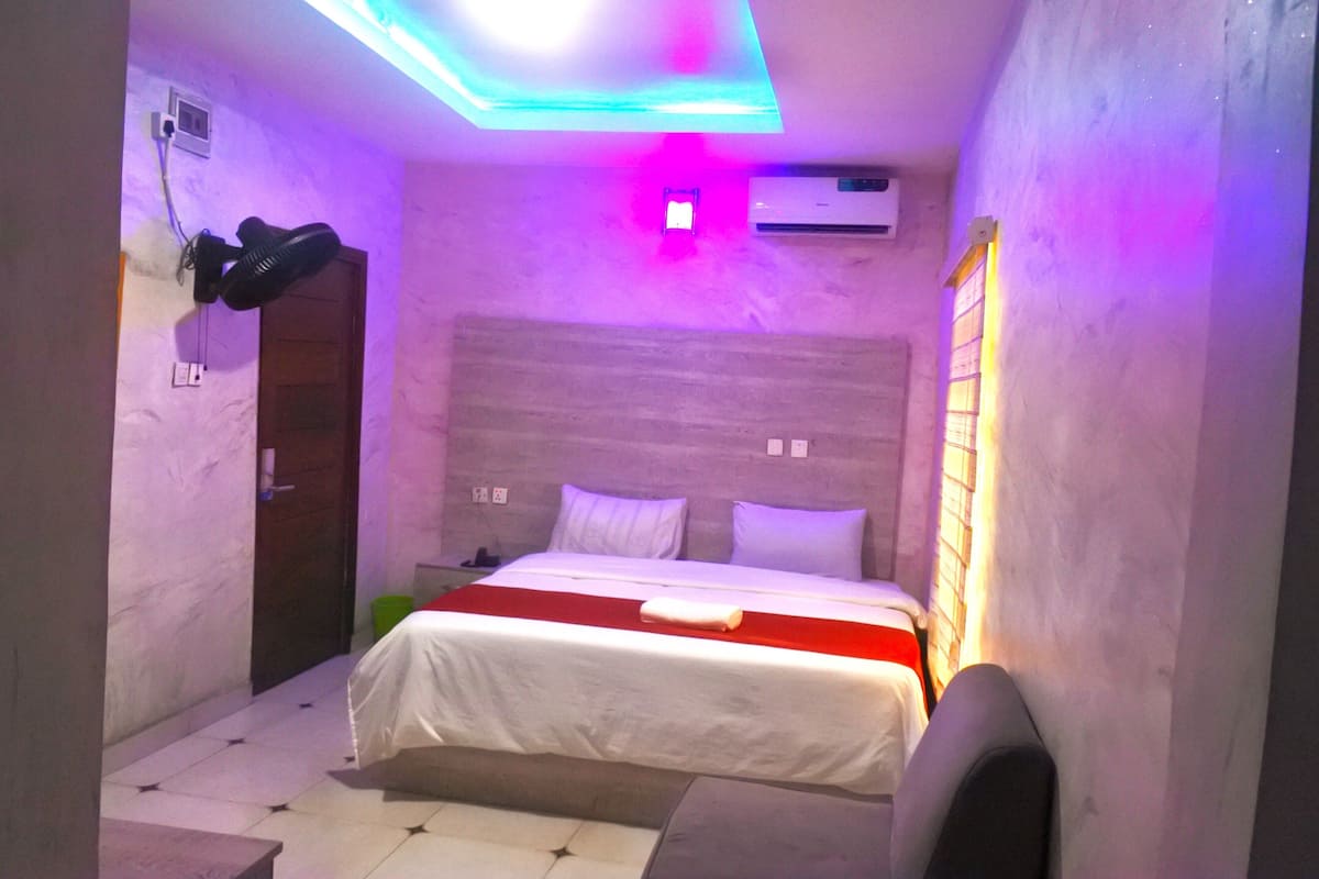 Un dormitorio con una cama, una mesita de noche, un ventilador de pared y un equipo de aire acondicionado.