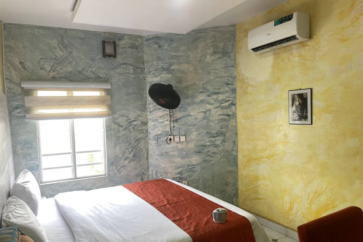 Un dormitorio con cama, ventana, aire acondicionado y ventilador de pared.