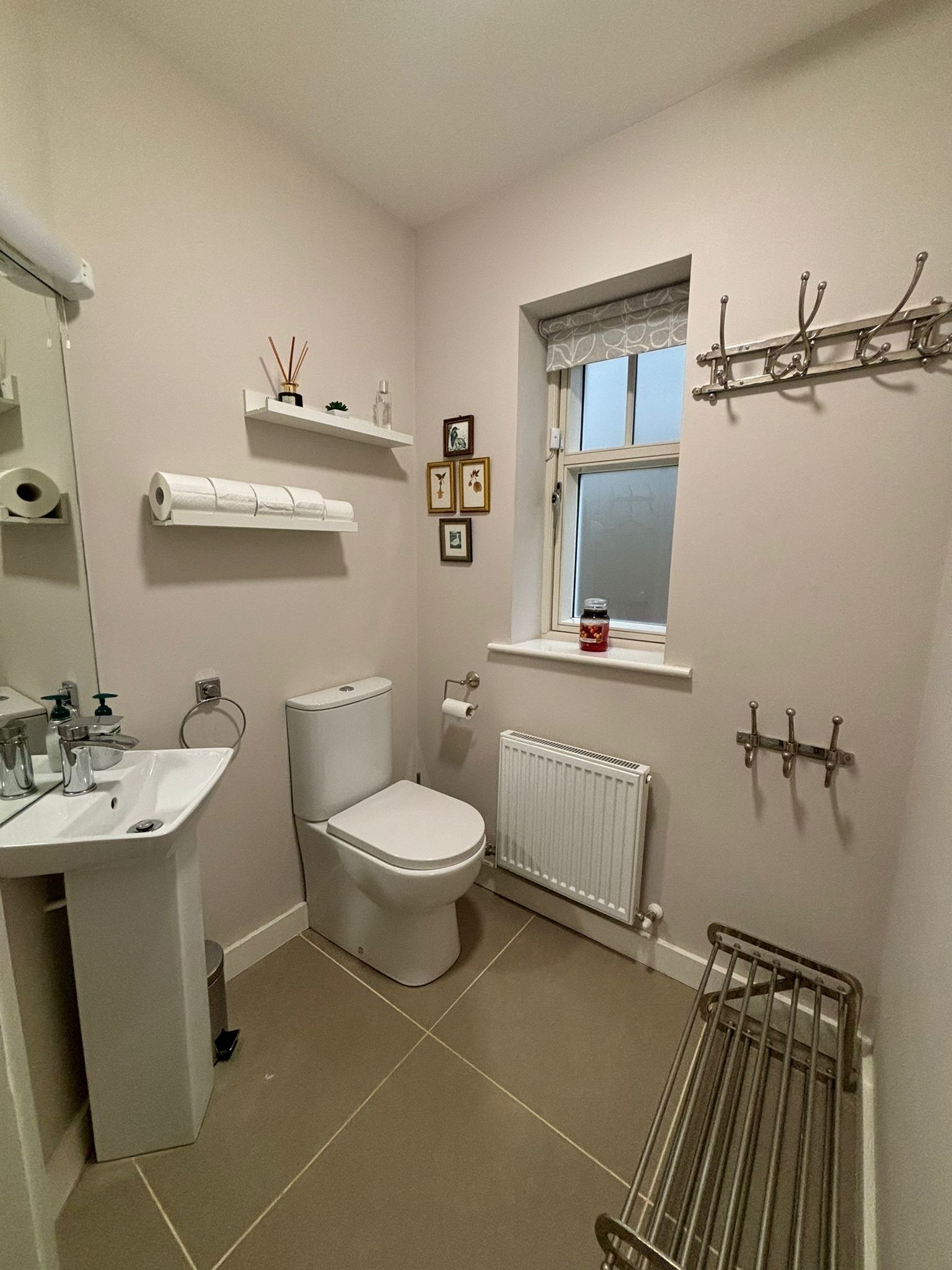 A small, clean bathroom with a toilet, sink, and radiator. There are picture frames on the wall and a window with a curtain.