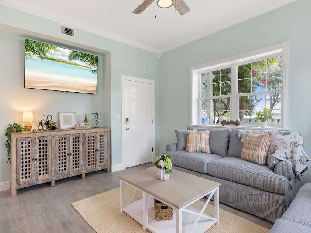 Una sala de estar con un sofá gris, una mesita de centro, un mueble con adornos, un televisor y una ventana con vistas a árboles.