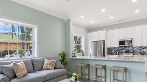 Eine moderne Küche mit weißen Schränken, einem Edelstahl-Kühlschrank und einer Mikrowelle. Ein graues Sofa mit gemusterten Kissen und ein kleiner Tisch mit einem Blumenstrauß.