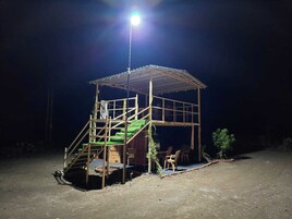 Una pequeña estructura de madera elevada con una escalera que conduce a una plataforma, rodeada de suelo de tierra.