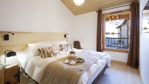 Un dormitorio con techo de madera, una cama grande con ropa blanca, una mesita de noche con lámpara y una ventana con vistas a edificios.