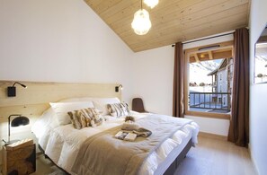 A bedroom with a wooden ceiling, a large bed with white bedding, a nightstand with a lamp, and a window with a view of buildings. - 3rd floor, fitness, swimming pool, sauna, hammam, balcony, tv, ski locker, 83m², Alpe d'Huez (Huez)