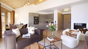 Una sala de estar moderna con sofá, sillón y mesa de centro, un comedor con mesa y sillas, y una cocina con gabinetes y electrodomésticos grises.
