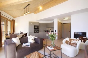 A modern living room with a sofa, armchair, and coffee table, a dining area with a table and chairs, and a kitchen with grey cabinets and appliances. - 3rd floor, fitness, swimming pool, sauna, hammam, balcony, tv, ski locker, 83m², Alpe d'Huez (Huez)