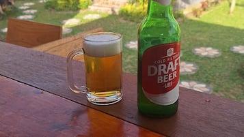 A glass of beer and a bottle of Draft Beer on a wooden table with a view of a wooden house in the background.