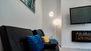 A modern living room with a black sofa, blue and mustard cushions, a flat-screen TV, a floor lamp, and a fireplace. - House - Sleeps 7 - Parking - Garden - Near Stn (Manchester)