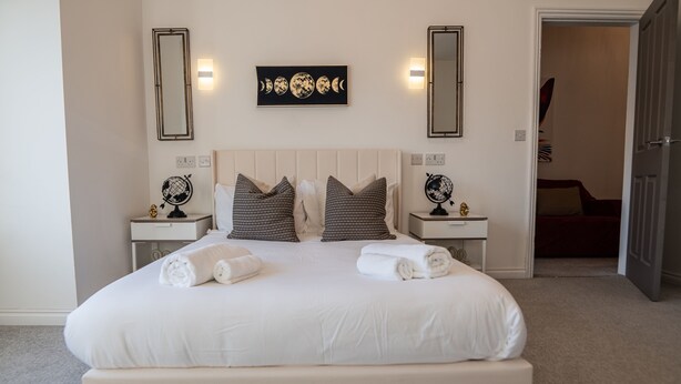 A neatly made bed with white linens and two pillows, a bedside table with a globe, and a wall-mounted artwork depicting moon phases. - House - Sleeps 7 - Parking - Garden - Near Stn (Manchester)