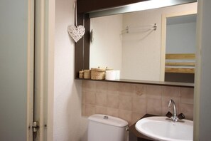 Ein Badezimmer mit Toilette, Waschbecken und Spiegel. An der Wand befindet sich eine herzförmige Dekoration und ein Regal mit Körben und Pflegeprodukten.