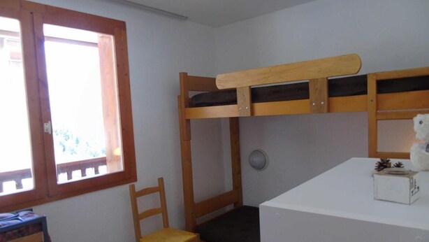 Uma cama de solteiro com estrutura de madeira, uma cadeira, uma pequena mesa com uma caixa e uma janela com moldura de madeira.