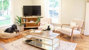 Een woonkamer met een bank, fauteuil en een hondenbed. Er is een televisie op een houten standaard, een glazen salontafel en een potplant.