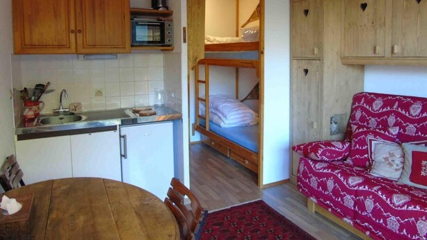 A compact room with a kitchenette, a bunk bed, a red patterned sofa, and wooden cabinets.