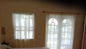 Ein Schlafzimmer mit einem großen Bett, zwei Fenstern mit durchscheinenden Vorhängen und Blick auf Grünfläche.