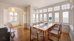 Um quarto espaçoso com uma grande mesa de jantar de madeira e cadeiras, um sofá de couro preto e vista para o mar através de grandes janelas com painéis de vidro decorativos.