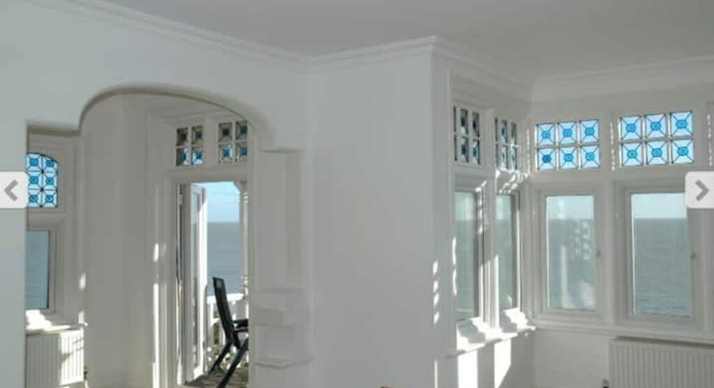 A room with white walls, a large window with blue-tinted panes, and a view of the sea.