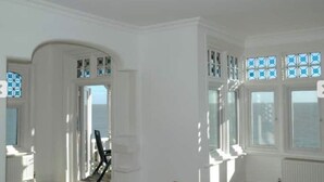 Quarto com paredes brancas, uma janela grande com vidros azulados e vista para o mar.
