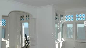 A room with white walls, a large window with blue-tinted panes, and a view of the sea.