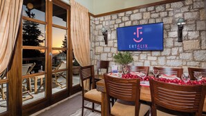A dining area with a stone wall, wooden chairs, a table set for four, and a TV mounted on the wall displaying 'ERTELIX'. - Sky Shades Villa in Arachova (Arachova)