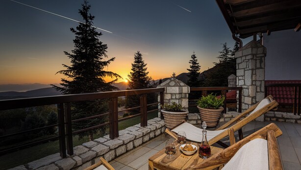 A wooden deck with lounge chairs, a table with drinks and snacks, and a view of the sunset over mountains. - Sky Shades Villa in Arachova (Arachova)