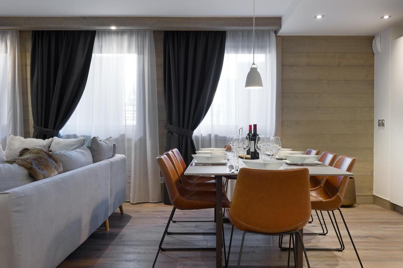 A modern living room with a dining area, featuring a sofa, dining table with chairs, and large windows with curtains.