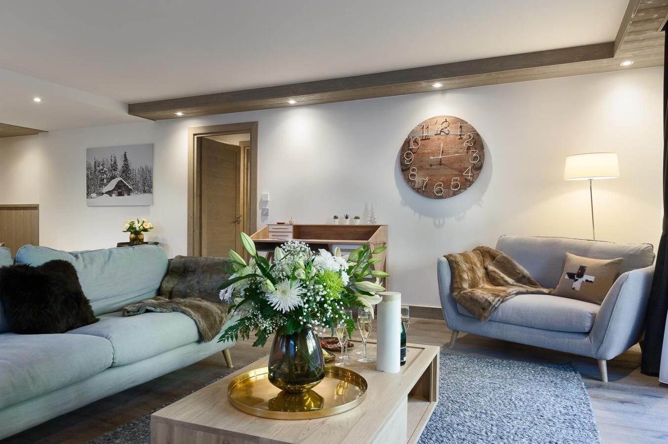 A modern living room with a light blue sofa, a grey sofa with a fur throw, a wooden coffee table with a vase of flowers, and a round wall clock.
