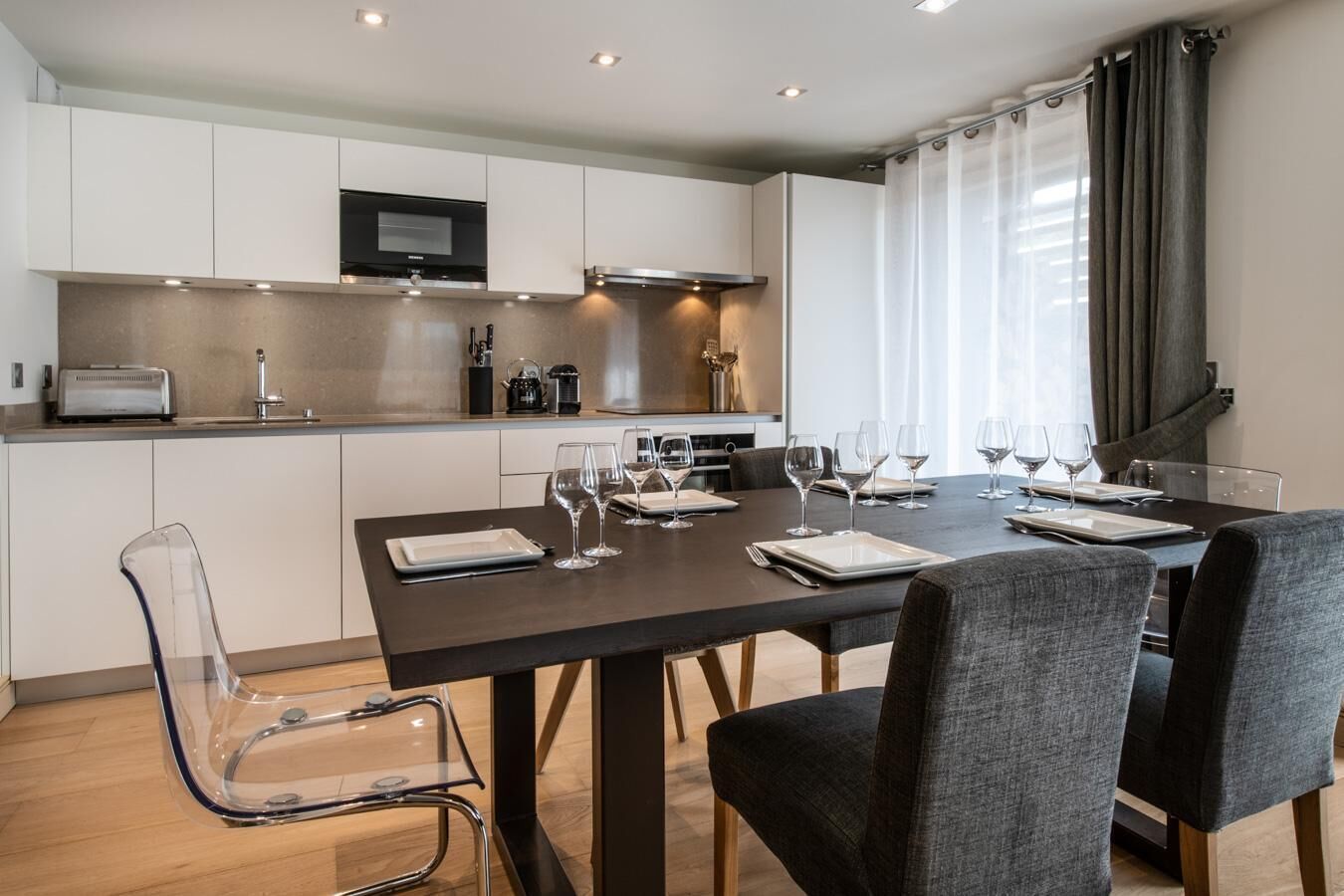 Una cocina moderna con armarios blancos, horno empotrado y microondas. Hay una mesa de comedor dispuesta con platos, vasos y cubiertos.
