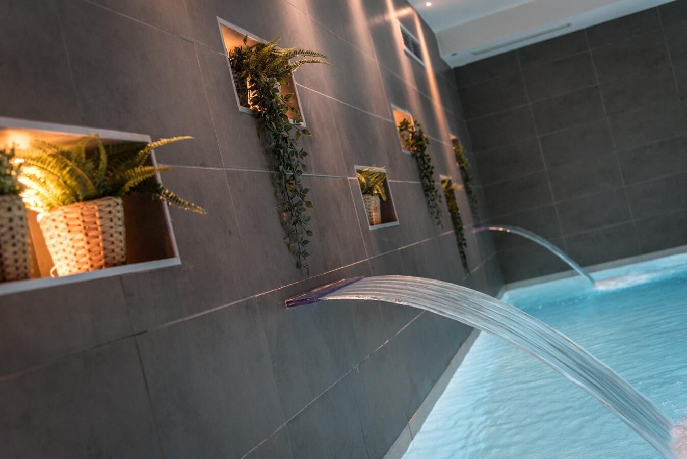 A modern bathroom with a large bathtub, grey tiled walls, and potted plants in built-in niches.