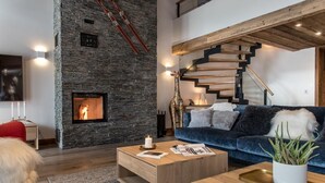 Una sala de estar moderna con chimenea de piedra, un sofá azul, una mesa de centro de madera y una escalera con vigas a la vista.