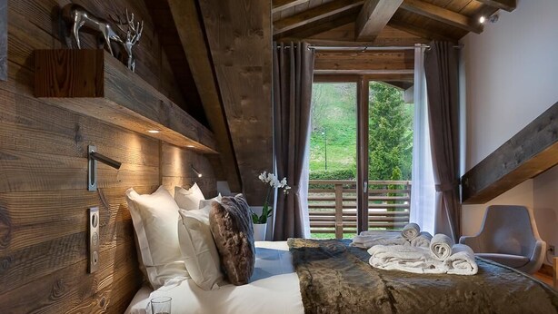 A wooden bedroom with a bed, pillows, a bedside table with a glass, and a view of greenery outside. - Wifi, resort center, 3rd floor, sauna, hammam, balcony, fireplace or stove, parking, tv, 158m² (Les Gets)