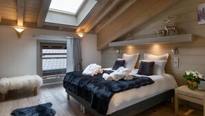 A bedroom with a skylight, a bed with white and dark pillows, a fluffy white chair, and a nightstand with a vase of flowers. - Wifi, resort center, 3rd floor, view mountain, sauna, hammam, terrace, balcony, fireplace or stove (Les Gets)