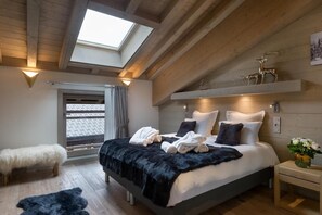 A bedroom with a skylight, a bed with white and dark pillows, a fluffy white chair, and a nightstand with a vase of flowers. - Wifi, resort center, 3rd floor, view mountain, sauna, hammam, terrace, balcony, fireplace or stove (Les Gets)