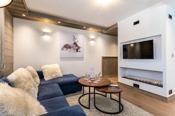 Un salón moderno con un sofá azul, mullidos cojines blancos, una mesa de centro redonda con vasos, una chimenea y un televisor montado en la pared.