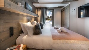 Une chambre d’hôtel moderne avec une tête de lit en bois, un grand lit recouvert de draps blancs, une couverture moelleuse et une vue sur un paysage enneigé par une porte ouverte.