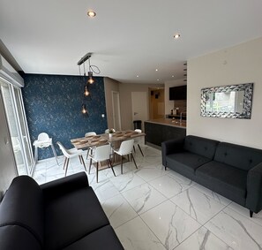 Una sala de estar moderna con un sofá oscuro, un comedor con una mesa y sillas de madera, y una cocina con encimera y armarios.
