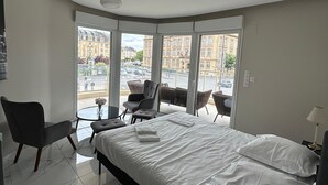 Habitación de hotel con cama, una silla, una mesita y vistas de la ciudad a través de amplios ventanales.