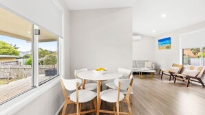A modern living room with a round table and white chairs, a grey sofa, and a view of the outdoors. - This is Livin' - Fire Pit - 3 Mins Walk to Beach (Culburra Beach)