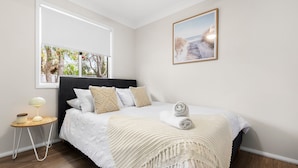 A bedroom with a large window, a bed with white bedding, a bedside table with a lamp, and a framed picture on the wall. - This is Livin' - Fire Pit - 3 Mins Walk to Beach (Culburra Beach)