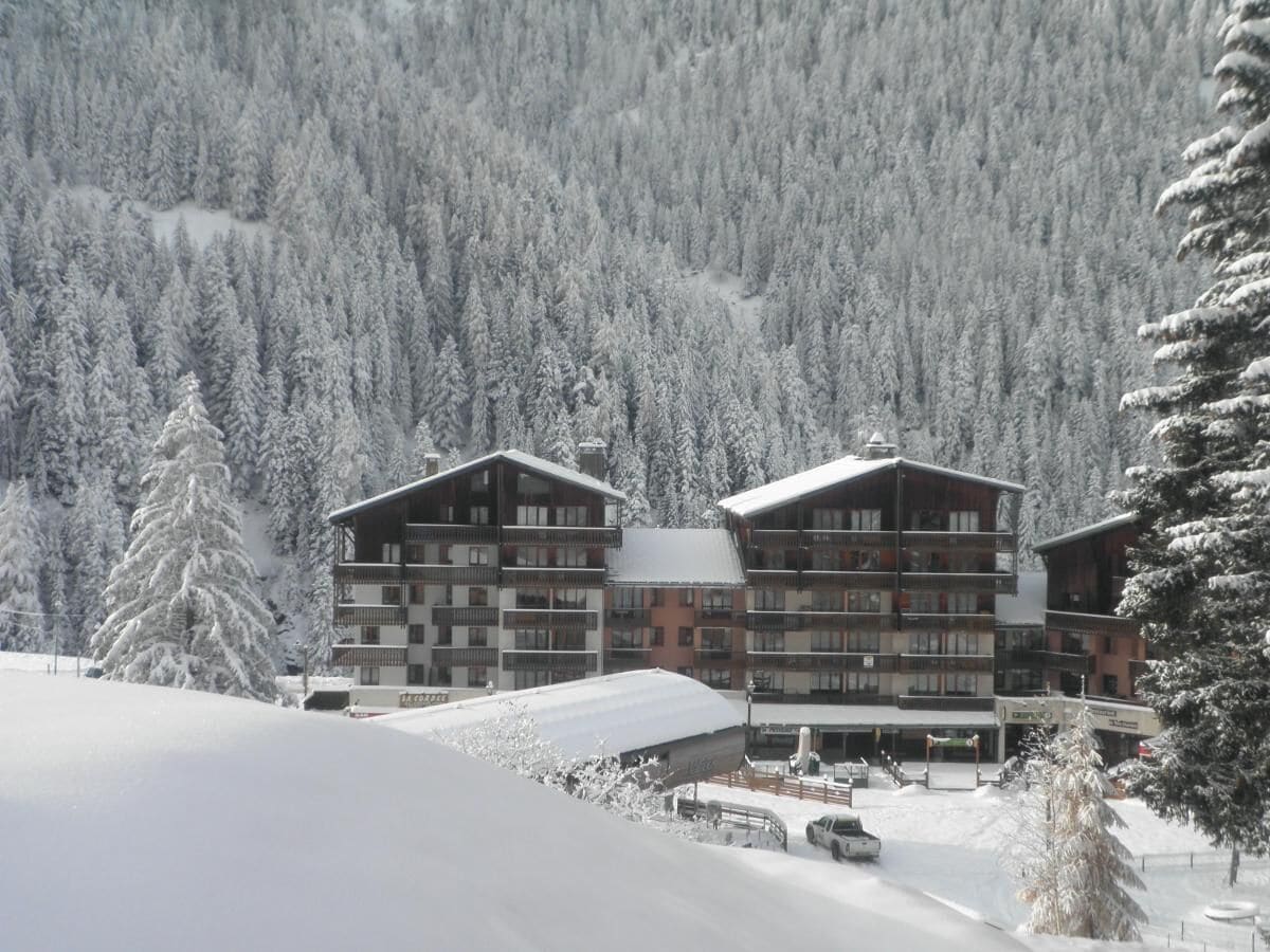 A multi-story hotel with balconies, surrounded by snow-covered trees and a mountainous backdrop.
