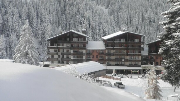 Un hotel de diversos pisos amb balcons, envoltat d'arbres coberts de neu i un paisatge de muntanyes.