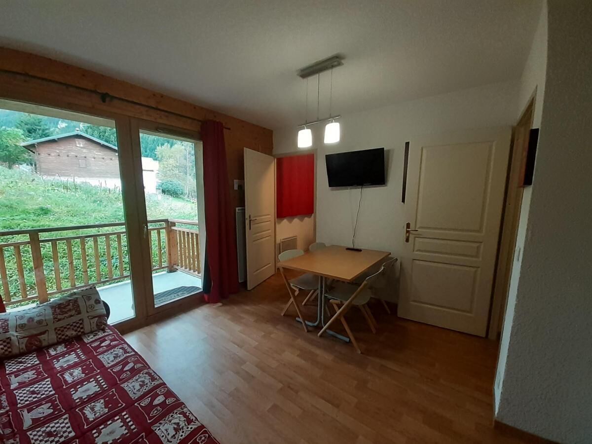 A room with a wooden floor, a table with chairs, a TV mounted on the wall, and a sliding glass door leading to a balcony.