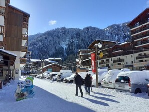 Eine verschneite Straße mit Gebäuden und geparkten Autos, bewölkter Himmel und einem Bergpanorama im Hintergrund.