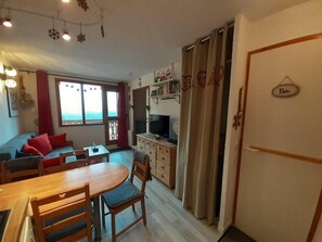Ein gemütliches Wohnzimmer mit einer Couch, einem Esstisch und einem Fernseher. Es gibt eine Küchenzeile mit einem Kühlschrank und einem Schrank.