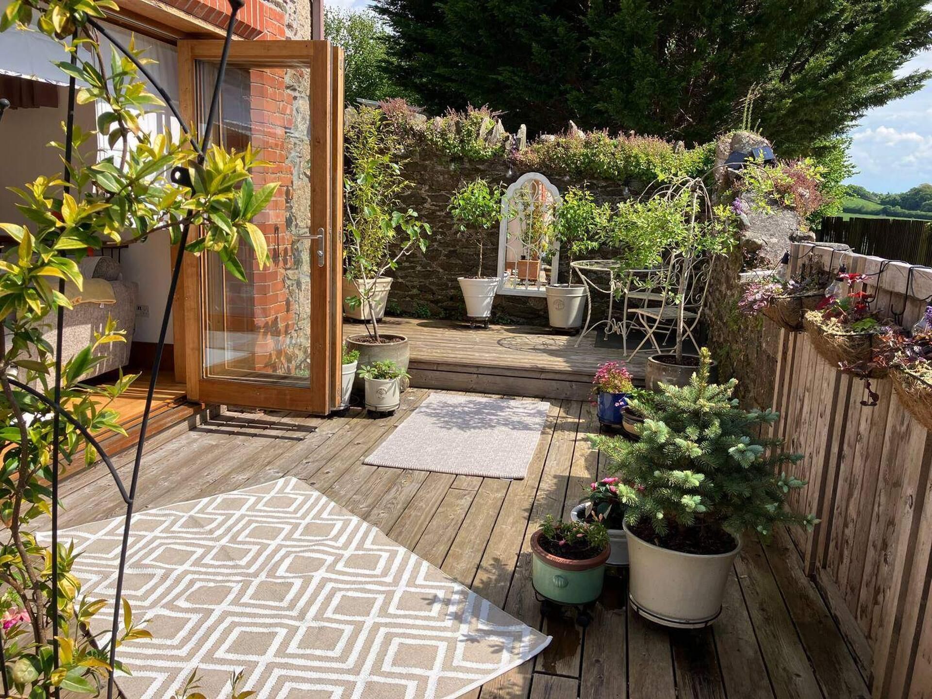 Eine Holzterrasse mit Blumentöpfen, einem gemusterten Teppich und einem Metallstuhl.