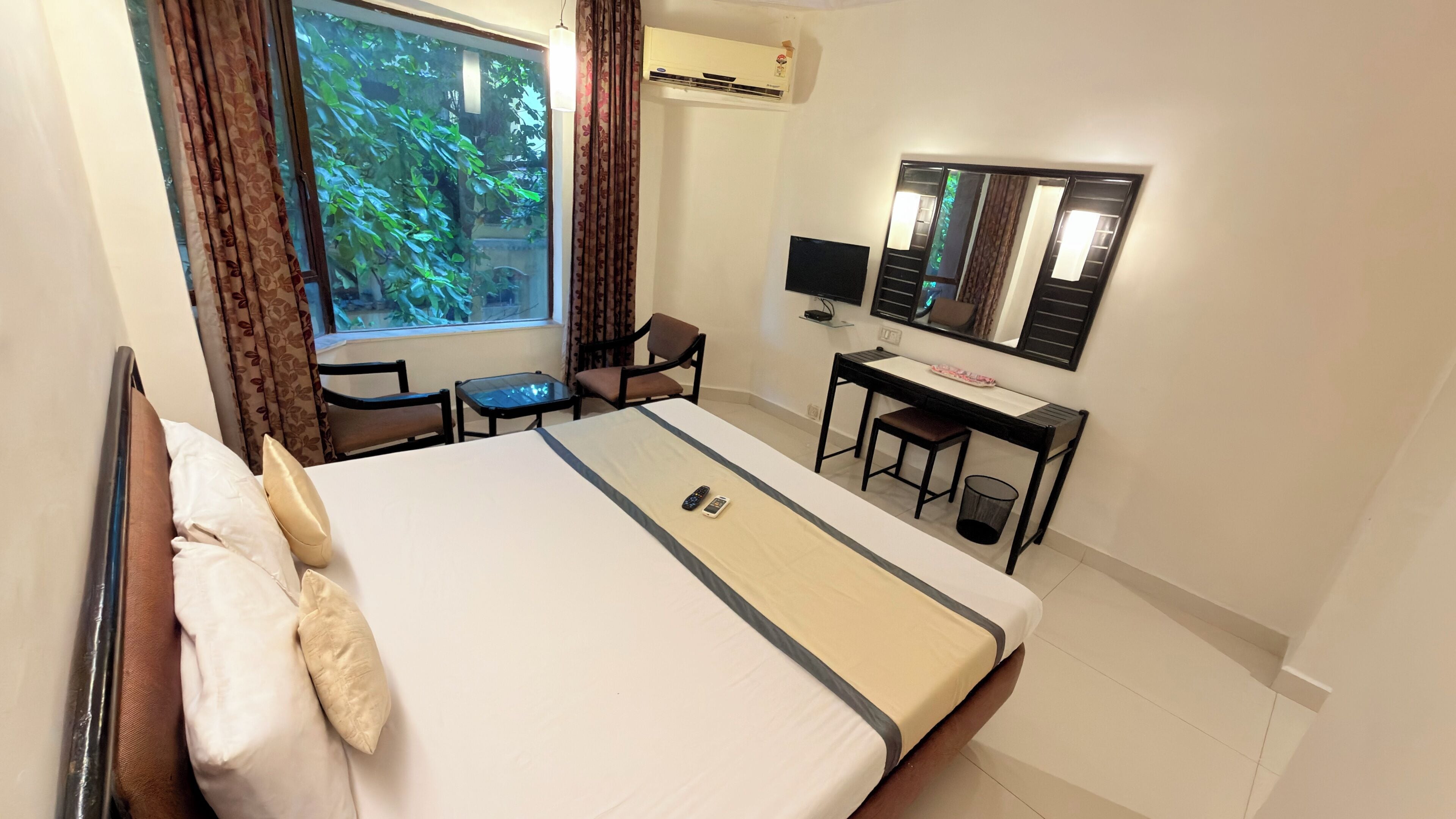 A hotel room with a bed, a desk, a chair, a TV, and an air conditioning unit.