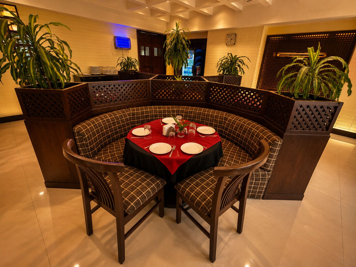 A round table with a red tablecloth, surrounded by checkered upholstered chairs, in a room with tiled flooring and potted plants.