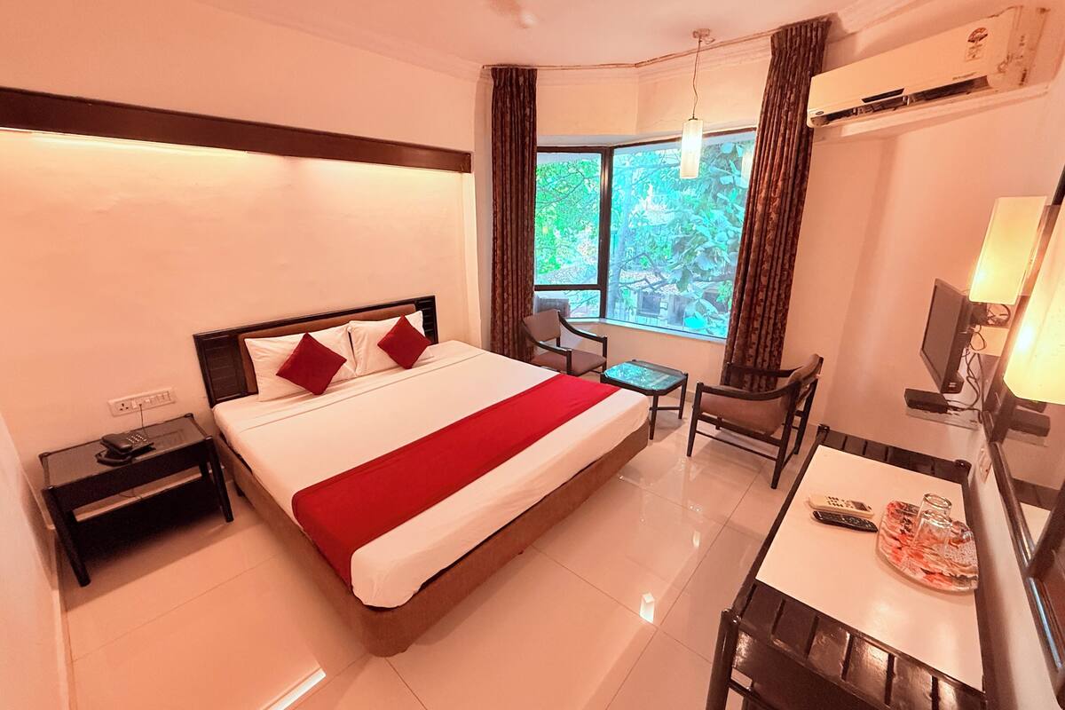 A hotel room with a bed, a desk, a chair, a TV, and a window with a view of trees.