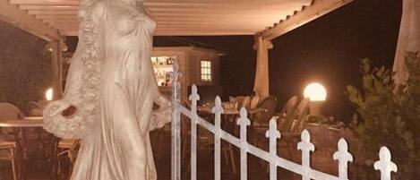 Estátua de mulher vestida de branco sob uma pérgula, com uma cerca branca ao fundo.
