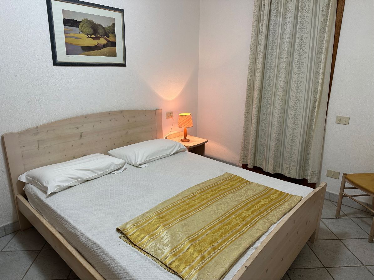 A neatly made bed with white linens and a yellow patterned throw, a bedside lamp, a framed painting on the wall, and a window with curtains.