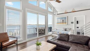 A spacious living room with a large window overlooking a body of water, a leather sofa, a dining area, and a staircase leading to an upper level.